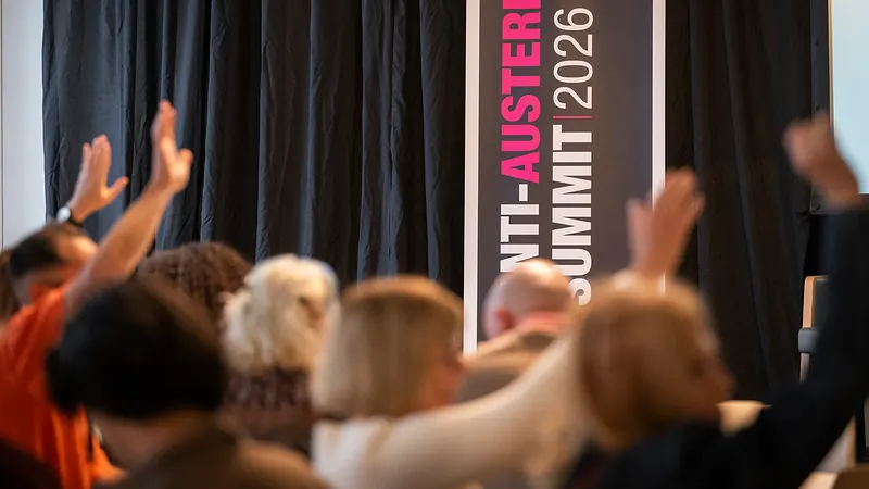 Delegates raise their hands in front of an Anti-Austerity Summit 2026 banner